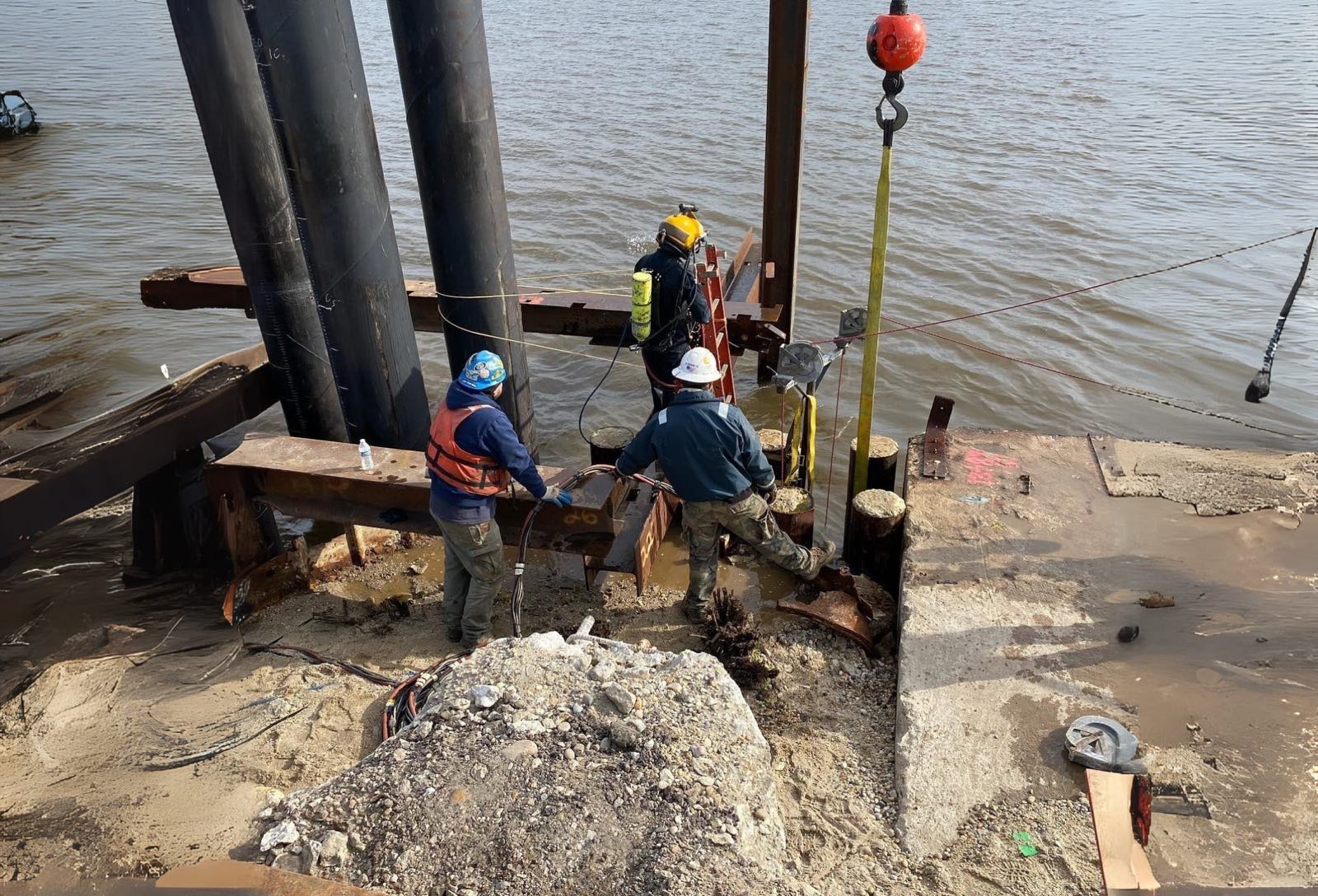 Diver entering the water at a tidal pile cluster while two crew members rig steel from the bank
